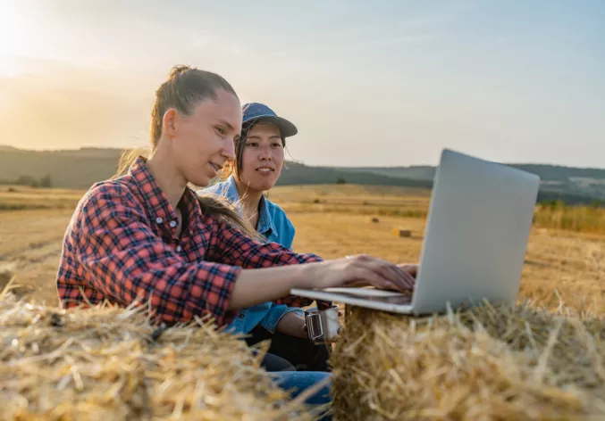 Two female farmers sitting on tractor trailer on haystacks and working on laptop together
